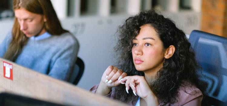 female with dark curly hair sitting at desk and looking pensively into the distance